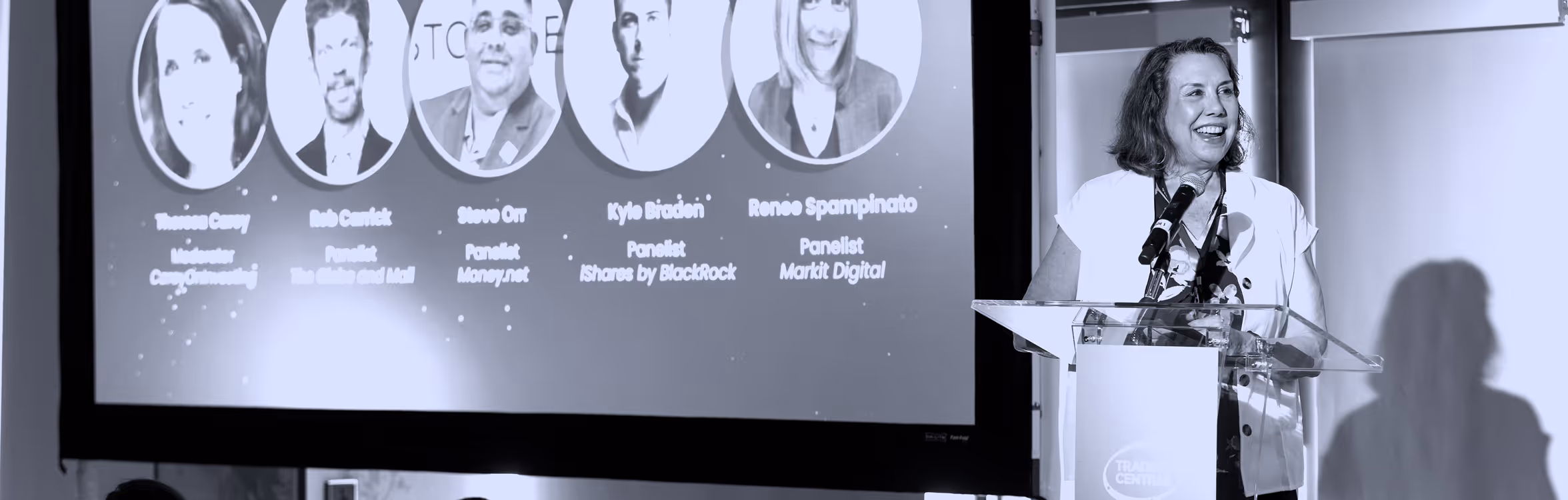 Speaker smiling and speaking at a podium with a panelist slide projected behind her.