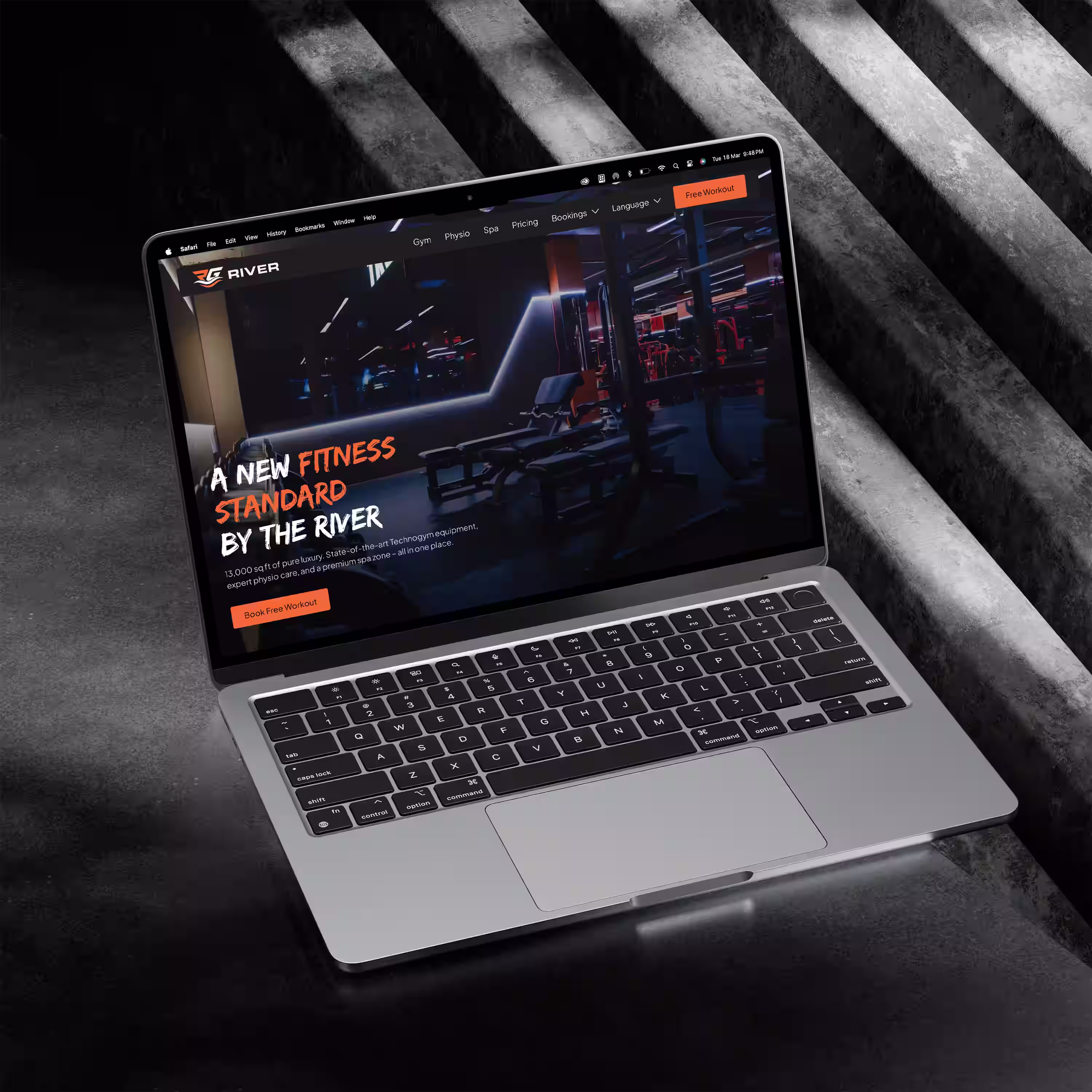 Open laptop on concrete floor displaying a fitness website with gym equipment and the text 'A New Fitness Standard by The River.'