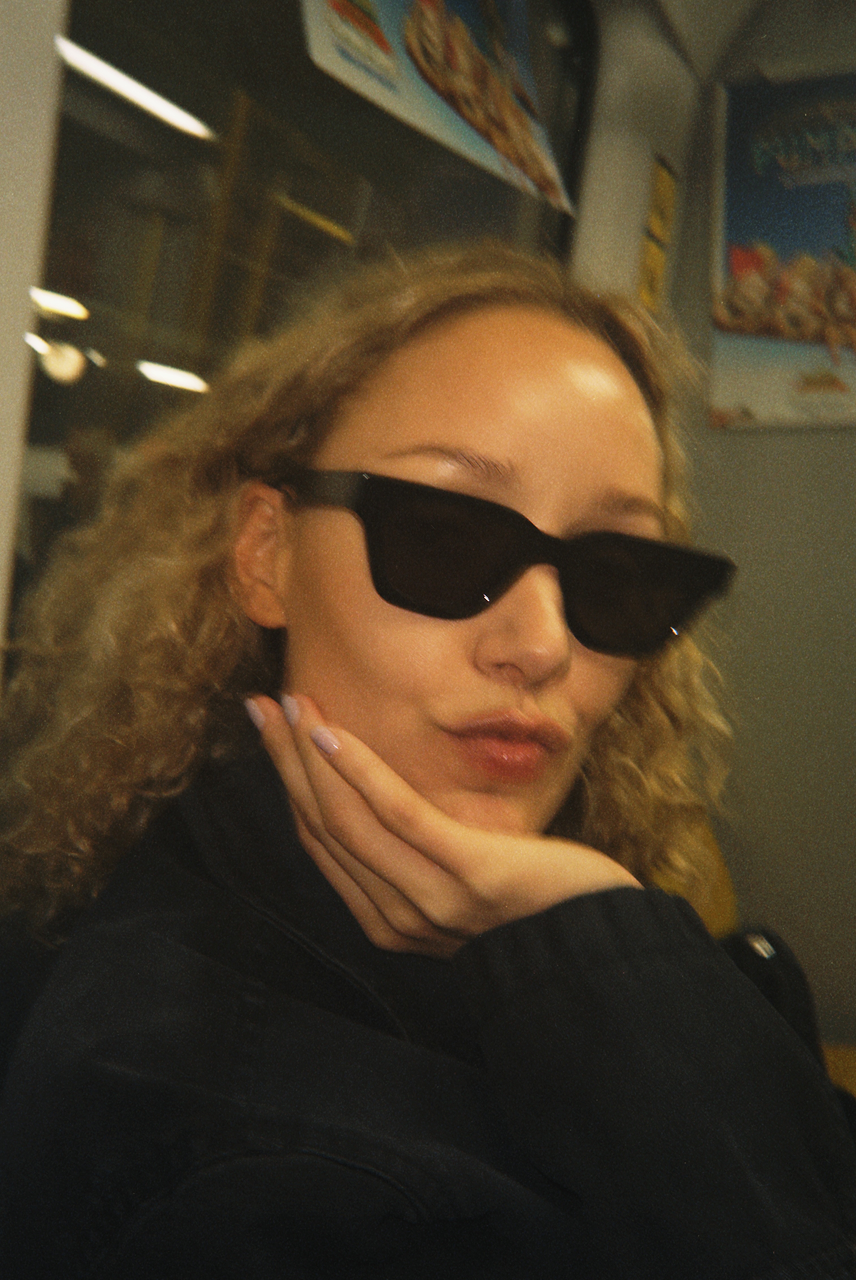 Person with curly blond hair wearing black sunglasses and a dark jacket, resting their face on their hand in an indoor setting.