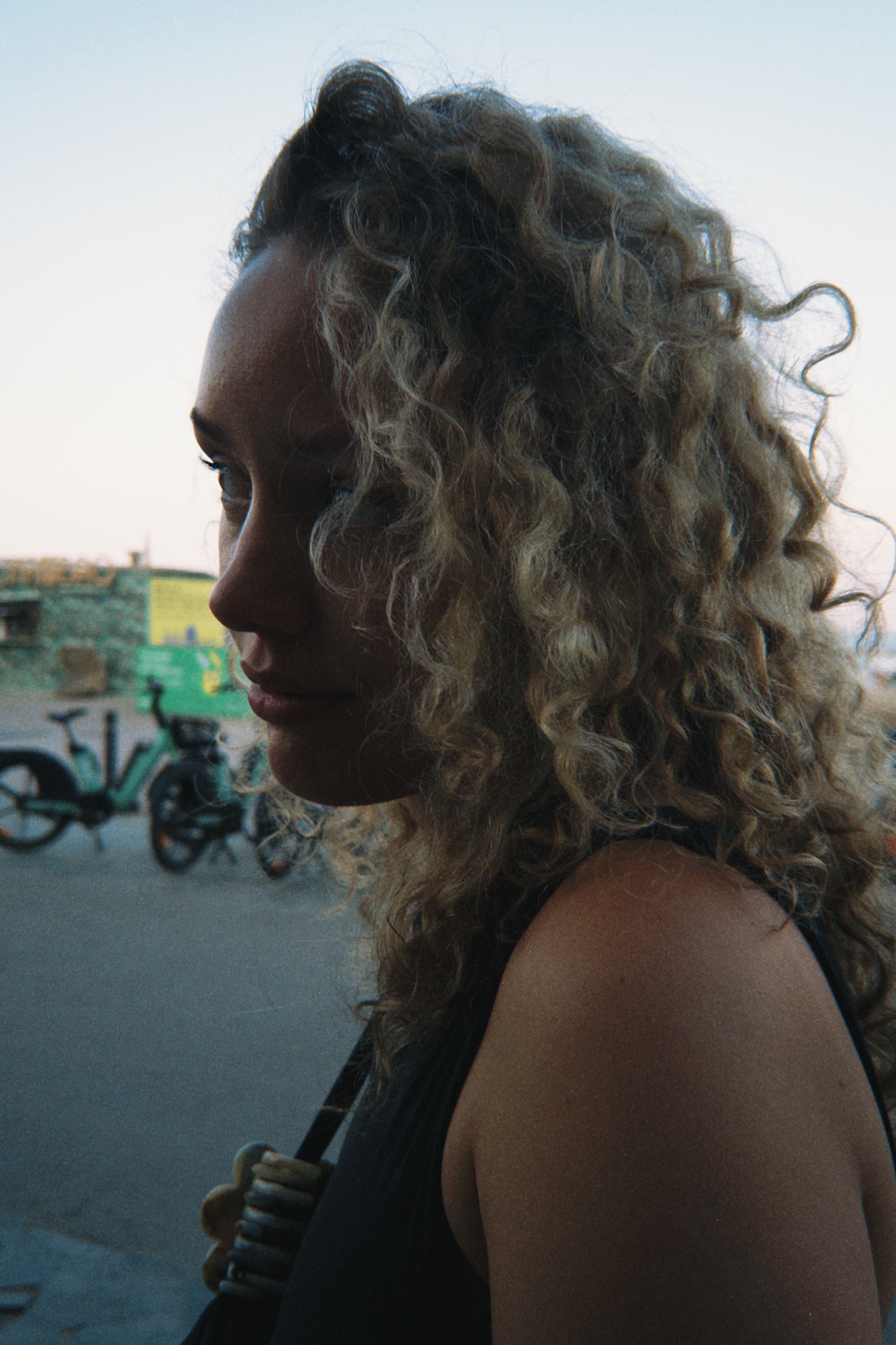 Side profile of a woman with curly blonde hair standing outdoors with bicycles in the blurred background.