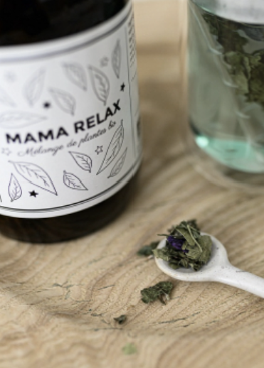 Close-up of a 'MAMA RELAX' jar, dried herbs on a white spoon, and a glass with herbs on a wooden surface.