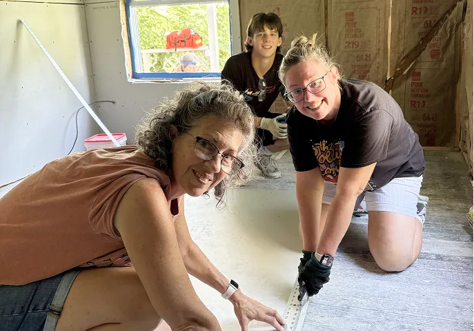 Three people kneeling on a floor inside a building under construction, smiling at the camera and working together.