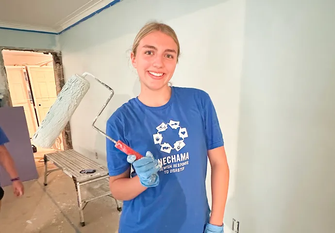 Smiling young woman wearing a blue t-shirt and gloves holding a paint roller in a room being painted.