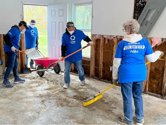 Volunteers wearing blue shirts and face masks cleaning a room with exposed wall insulation using brooms and a wheelbarrow.
