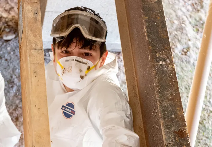 Person wearing protective white coveralls, safety goggles on forehead, and a dust mask, working between wooden and rusty metal beams.