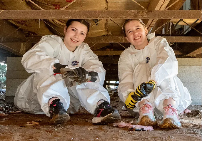 Two smiling women in white protective suits and gloves sitting on dirt under a wooden structure.