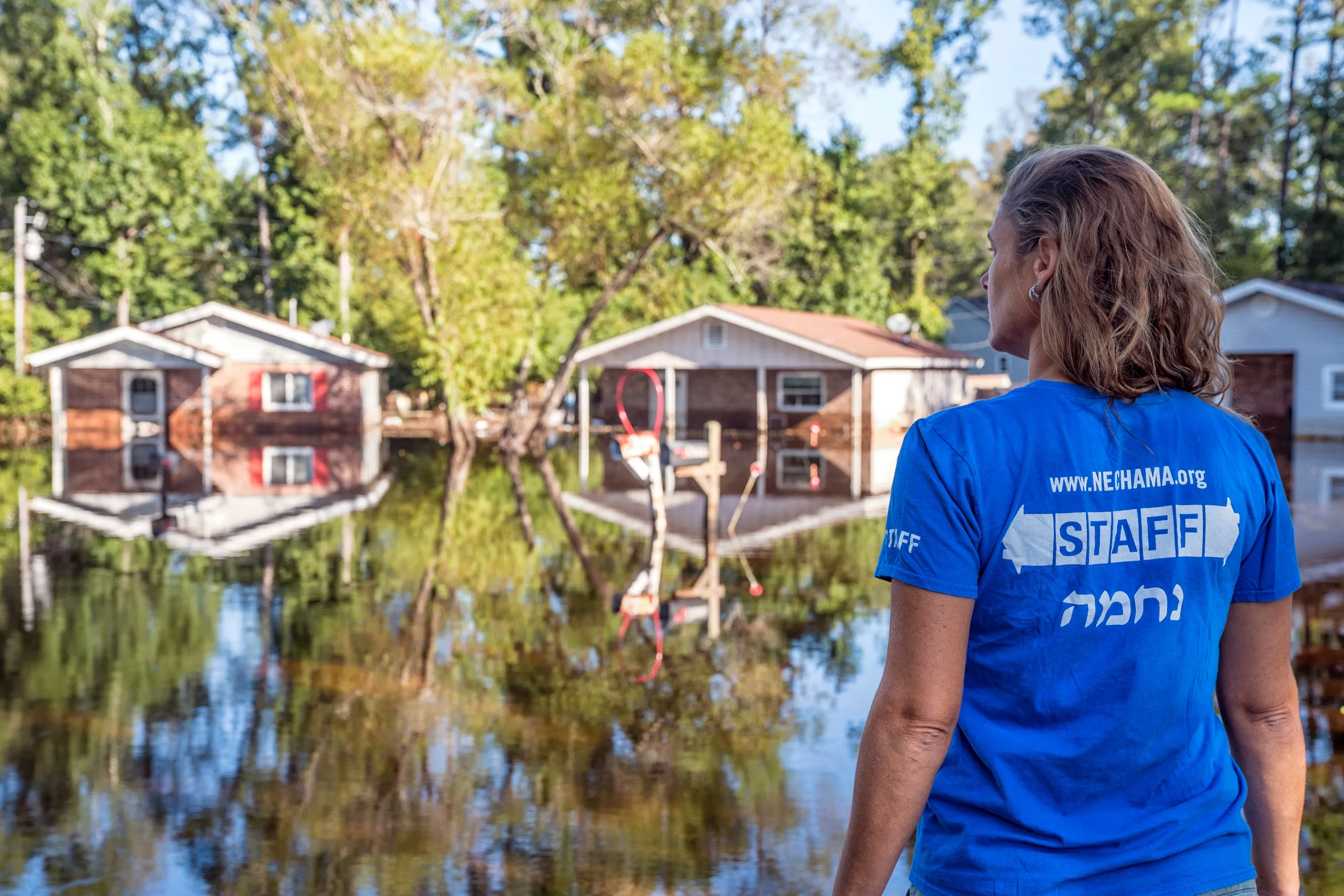 Person wearing a blue staff shirt stands looking at houses partially submerged in floodwater.