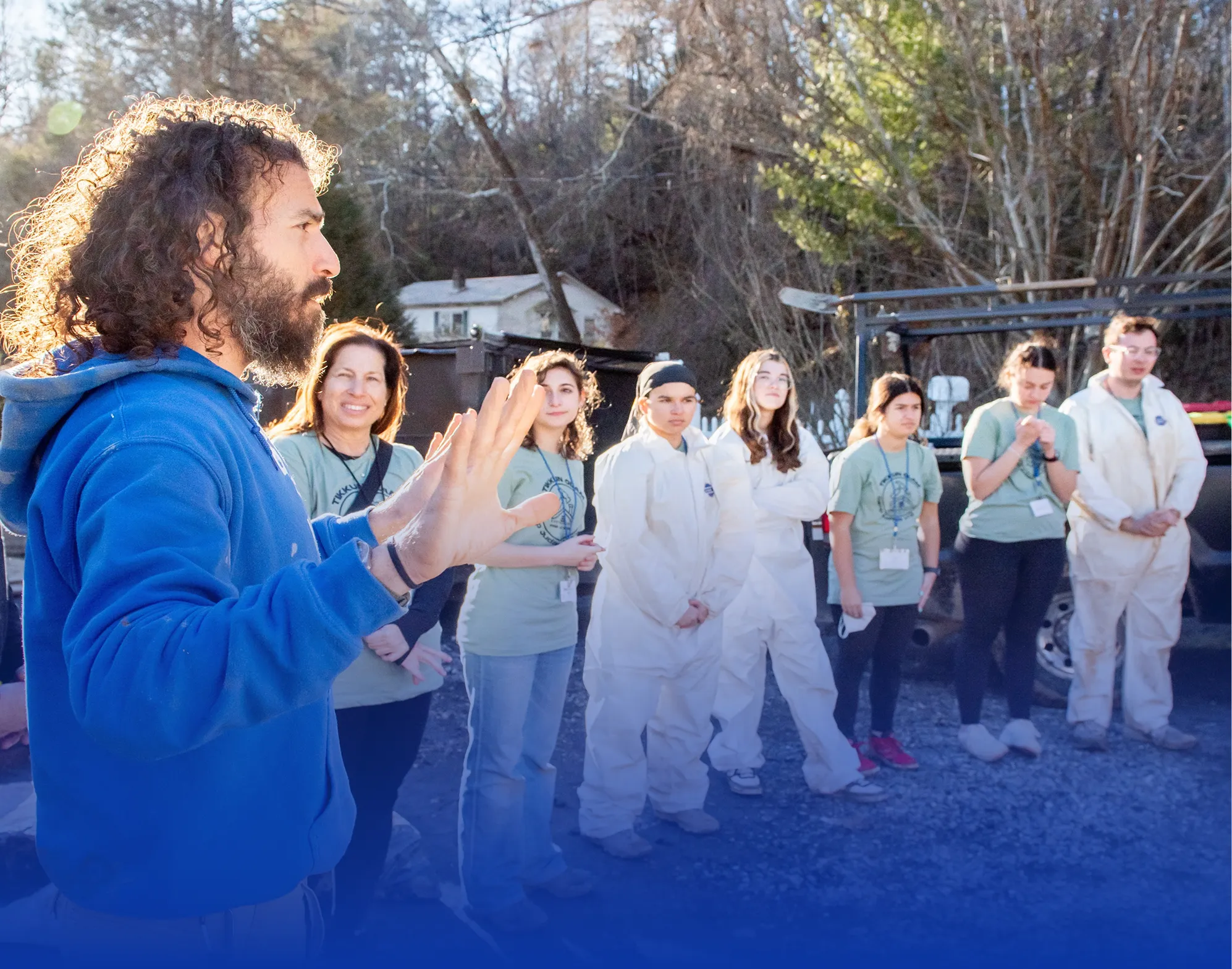 Man in a blue hoodie speaking and gesturing to a group of people, some wearing light green shirts and others in white protective suits, standing outdoors on gravel with trees in the background.