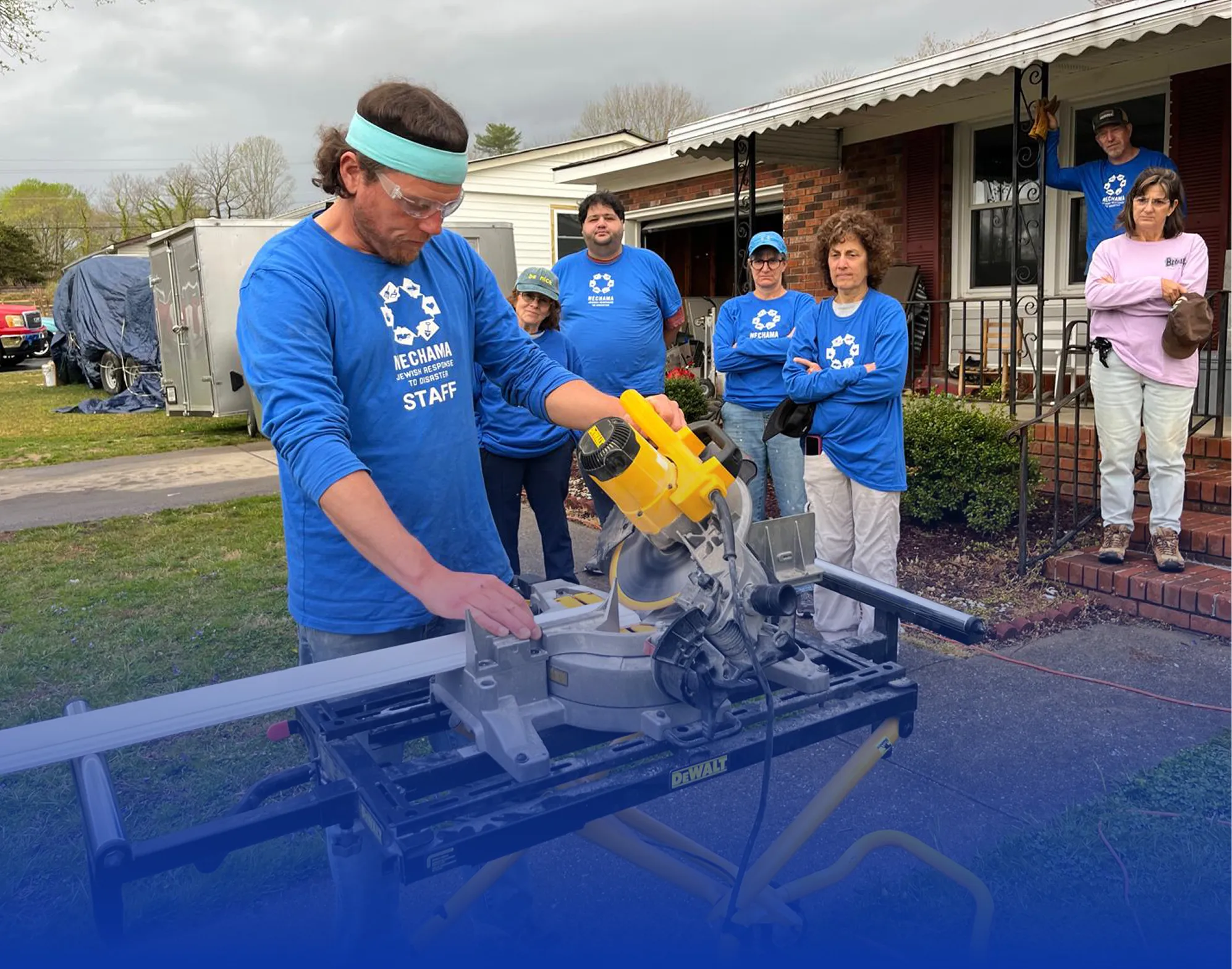 Man wearing safety glasses and a headband operates a miter saw outdoors while five people in blue and pink shirts watch near a house.