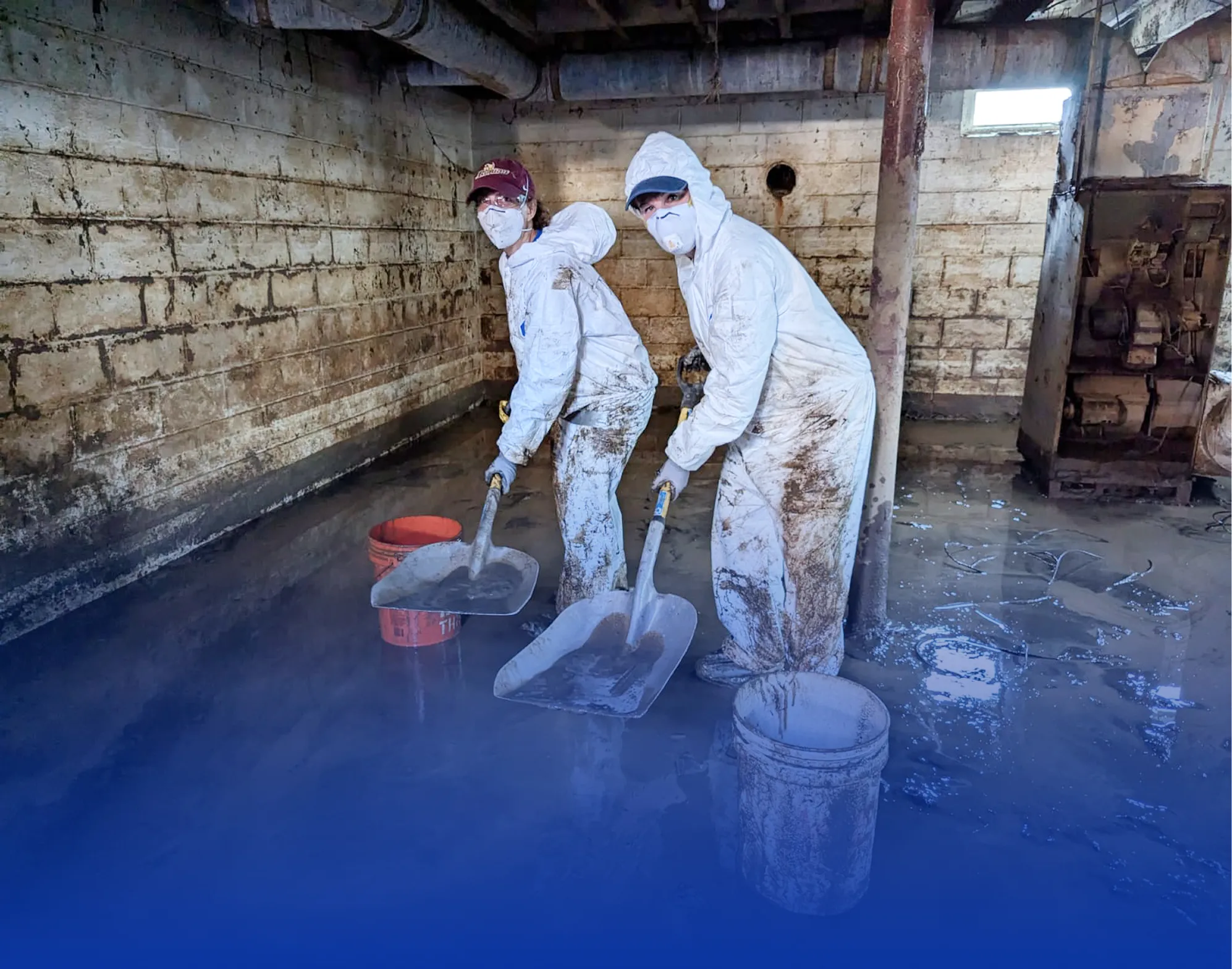 Two workers in white protective suits and masks shoveling muddy water inside a basement or flooded room.