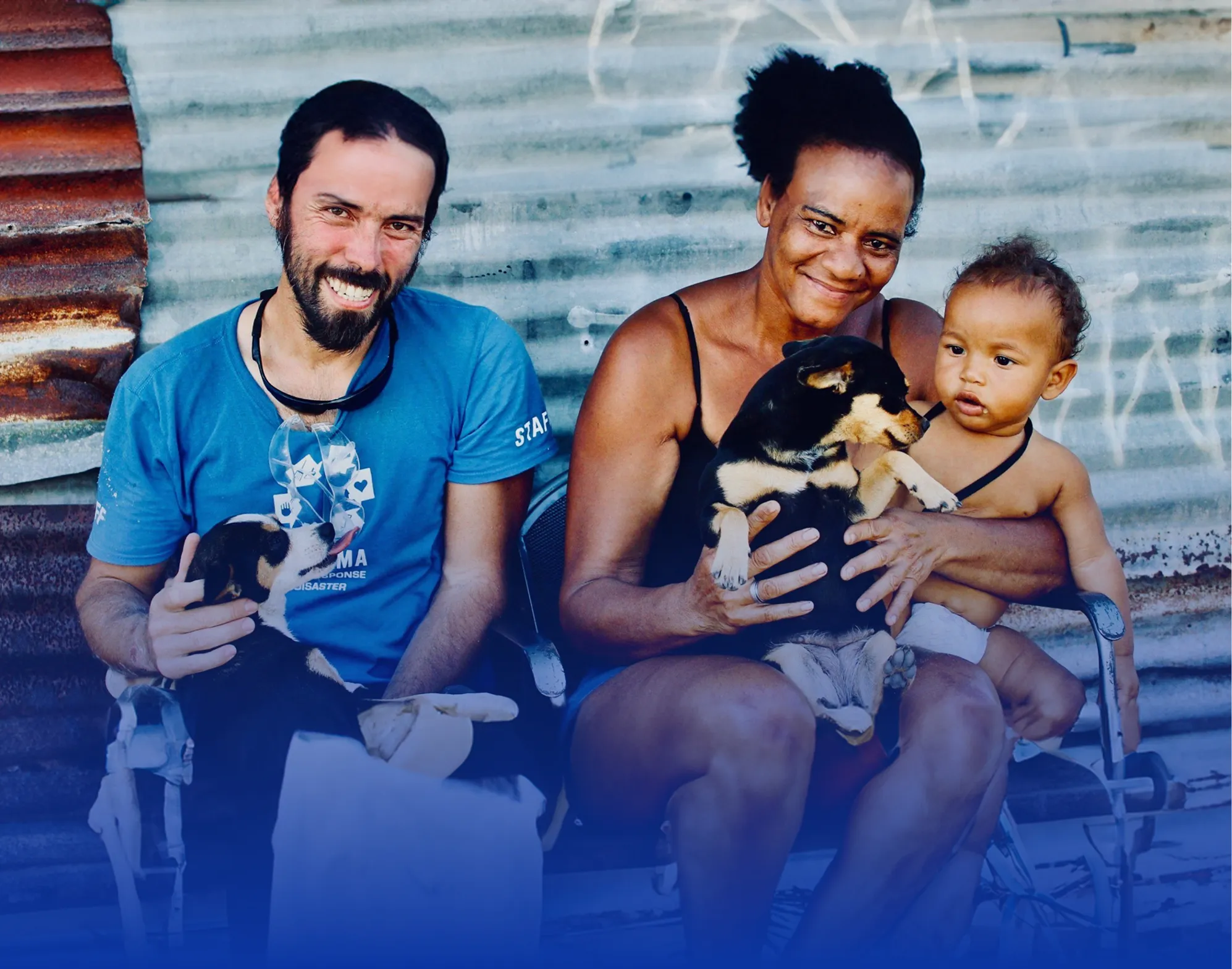 Smiling man and woman holding small dogs, with a toddler sitting on the woman’s lap against a corrugated metal background.