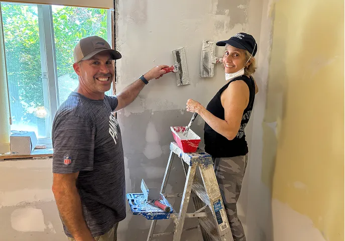 A man and a woman smiling while plastering a wall together inside a room under renovation, with a window and a ladder nearby.