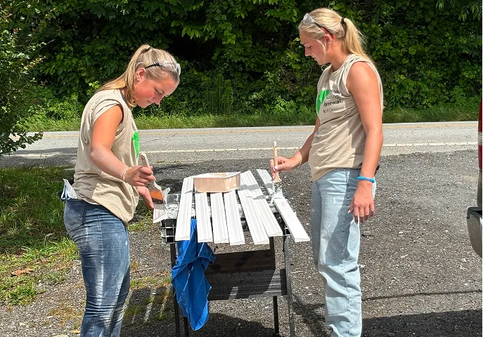 Two women painting wooden slats white outdoors on a sunny day.
