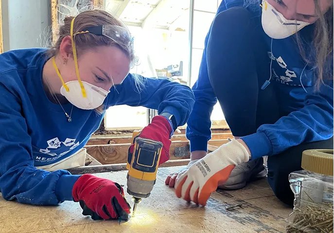 Two people wearing face masks and gloves using a power drill together while working on a wooden surface indoors.