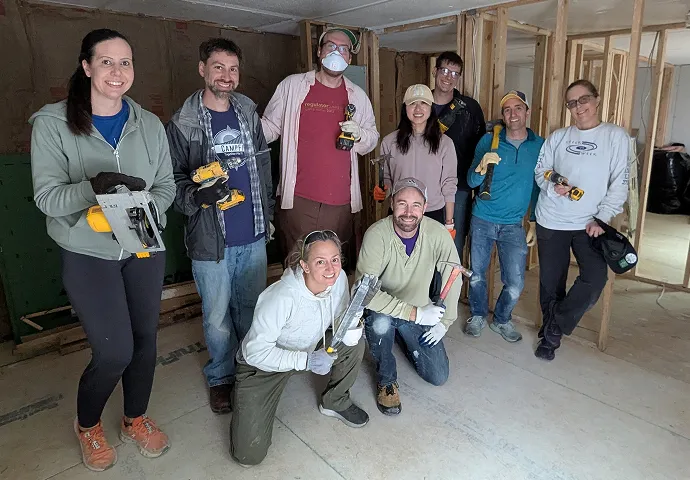 Group of nine people smiling indoors, holding construction tools, standing and kneeling in a partially framed room under renovation.