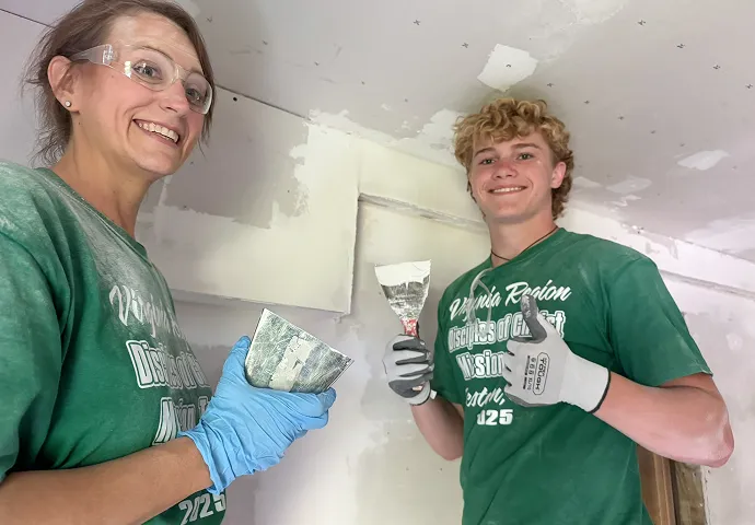 Two people wearing green shirts and gloves, smiling while applying joint compound on drywall using putty knives.