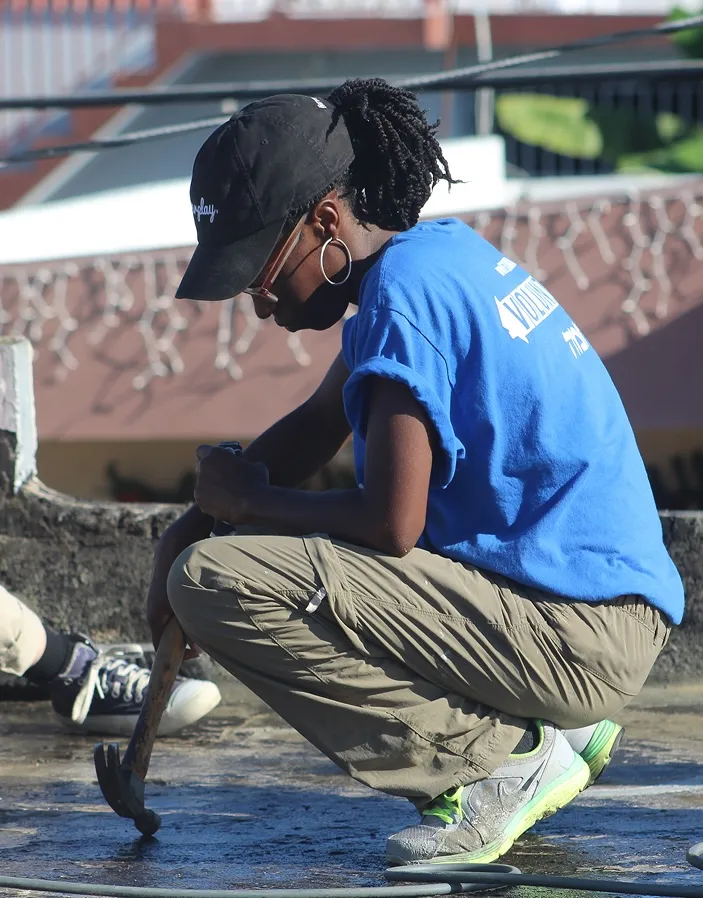 Person wearing a blue t-shirt, beige pants, and a black cap crouching and using a hammer on the ground outdoors.