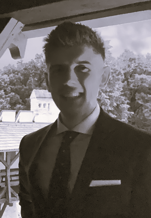 Prezes firmy, mężczyzna w garniturze i krawacie stoi na zewnątrz, w tle drzewa i dach budynku.