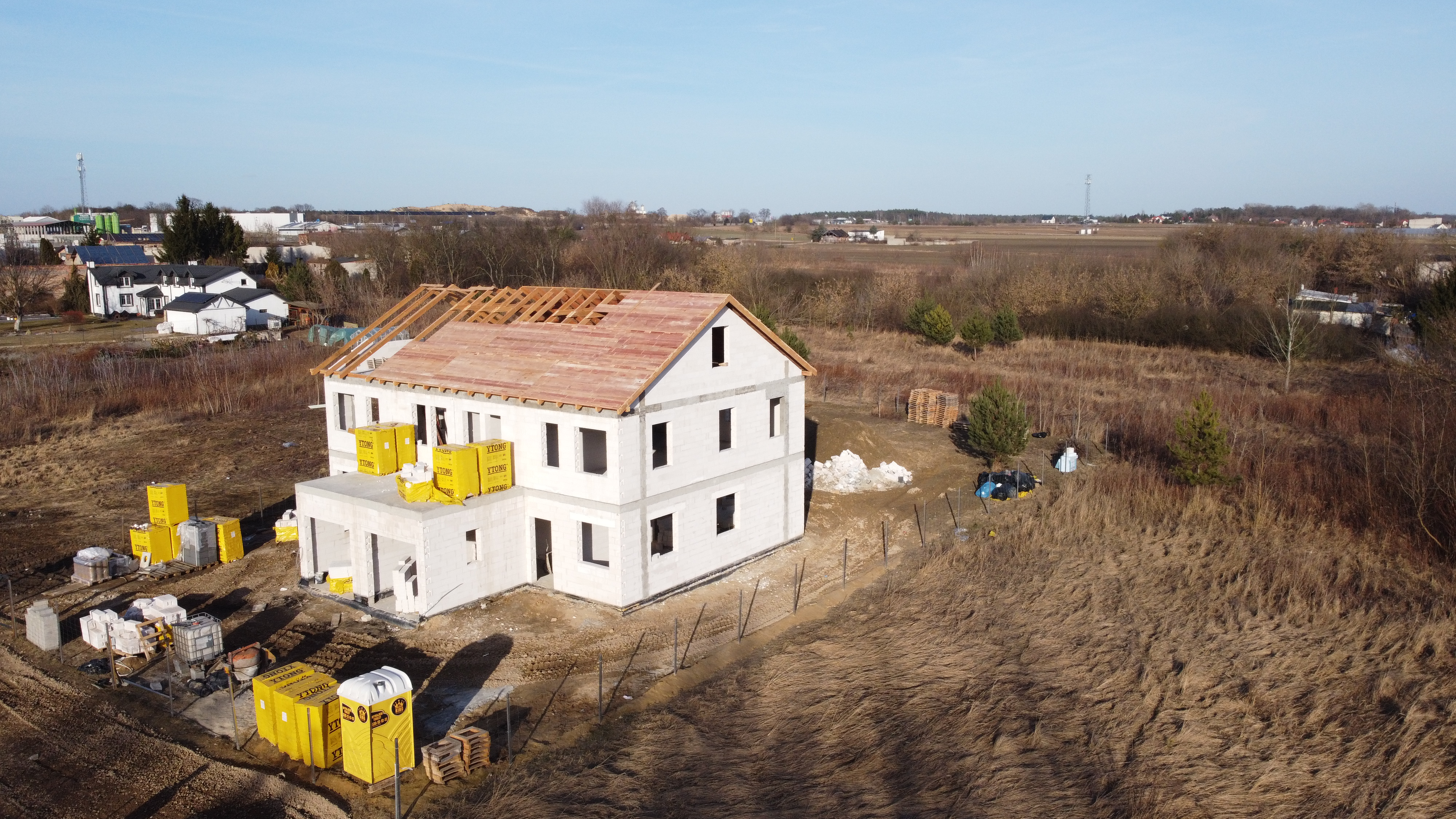 Dom w trakcie budowy na pustej działce z widokiem na pola i niską zabudowę w tle.