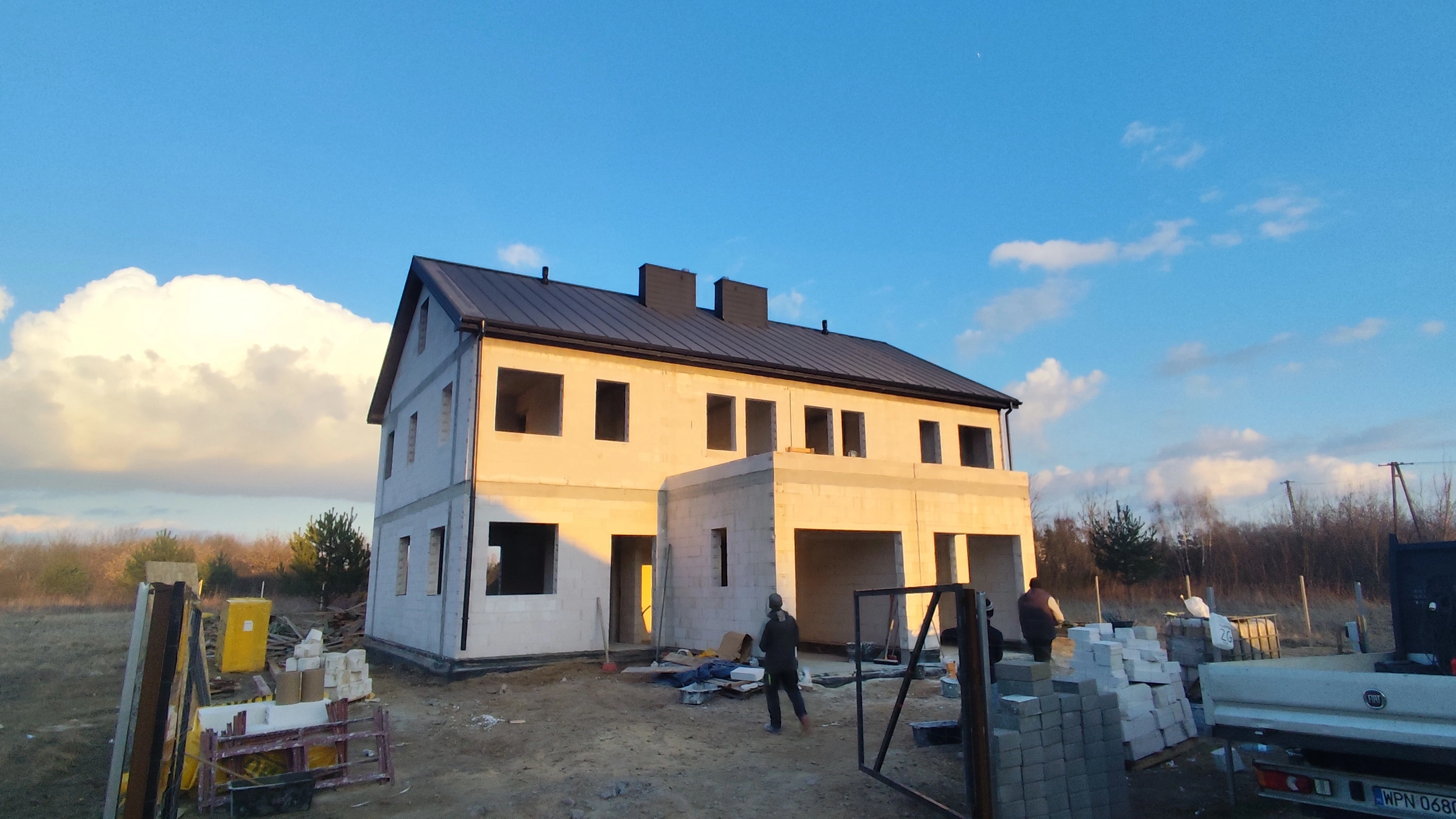 Dom w trakcie budowy na nieutwardzonej działce z dwoma osobami przy stosach materiałów budowlanych.