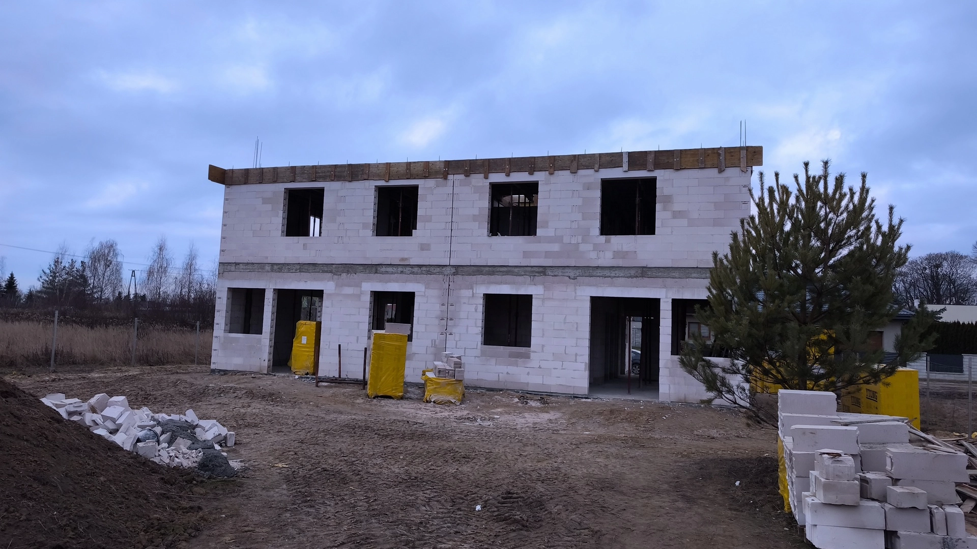 Niedokończony budynek mieszkalny z bloczków betonowych na placu budowy, z żółtymi materiałami budowlanymi na zewnątrz.
