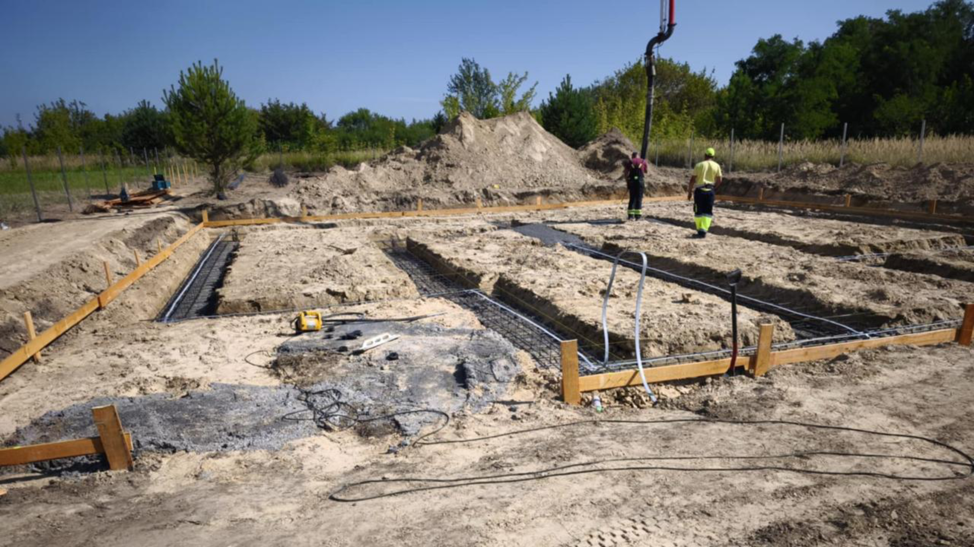 Plac budowy z przygotowanym fundamentem, drewnianymi szalunkami i pracownikami przy wylewaniu betonu.