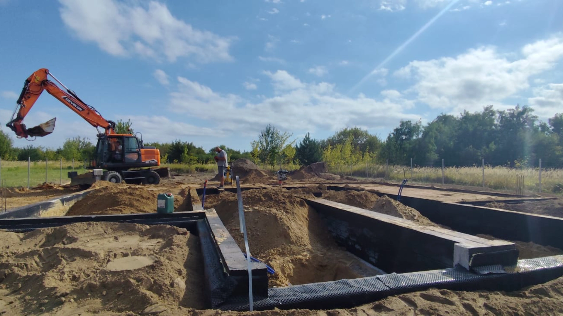 Plac budowy z widocznym wykopem fundamentowym, koparką oraz pracownikiem na tle drzew i nieba.
