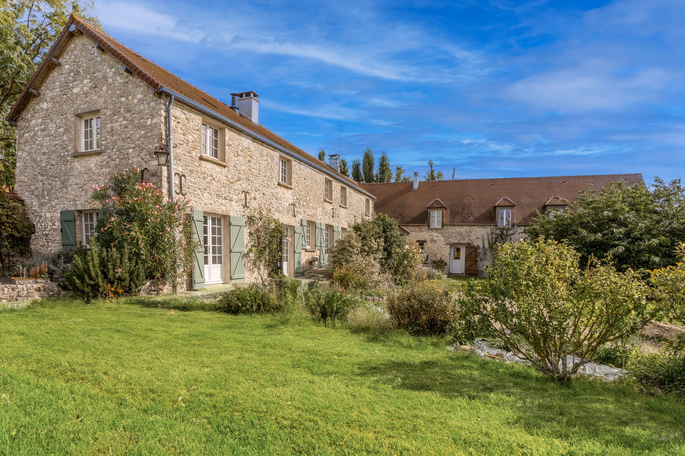 Stone countryside house with green shutters, surrounded by lush garden and a large green lawn under a blue sky.