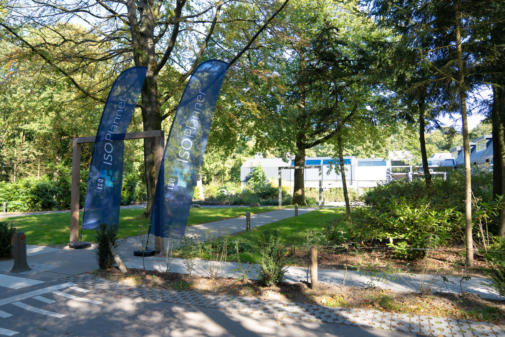 Two ISOPlanner™ flags stand by a wooden archway on a garden path, with the conference building visible among trees.