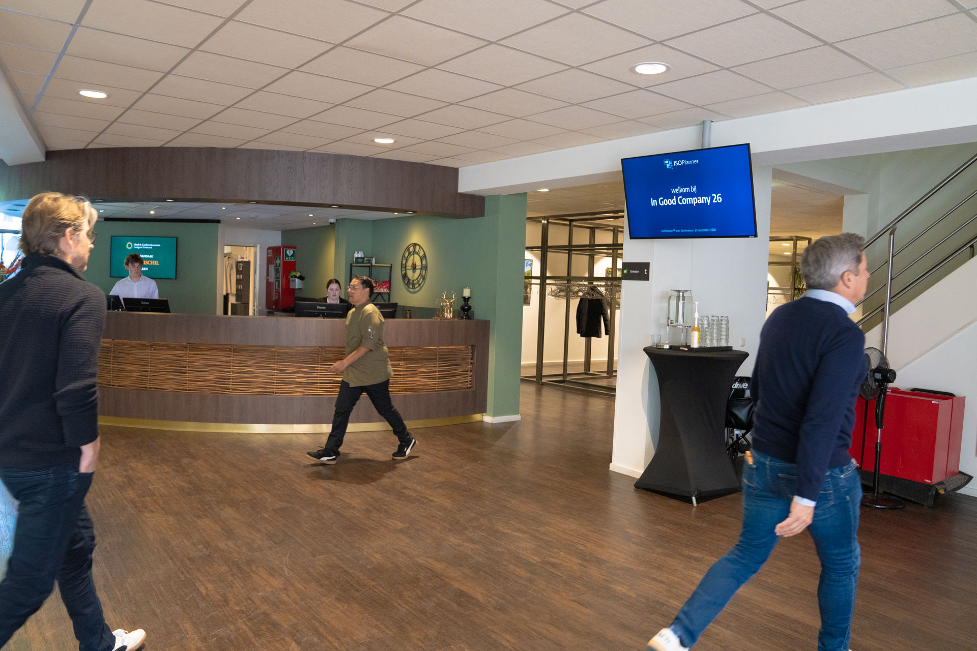 Three people cross a hotel lobby past the reception desk, with a screen reading welkom bij In Good Company 26.