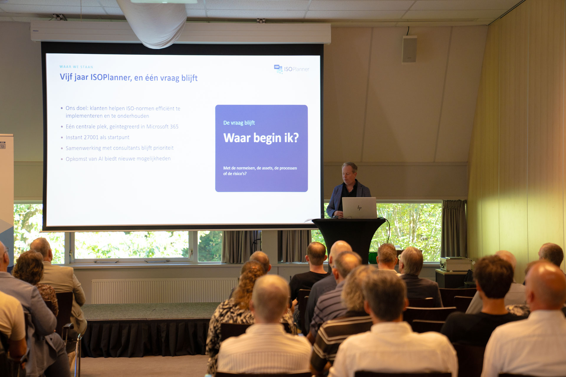 A speaker stands at a podium beside a screen reading Vijf jaar ISOPlanner™, en één vraag blijft, addressing the room.
