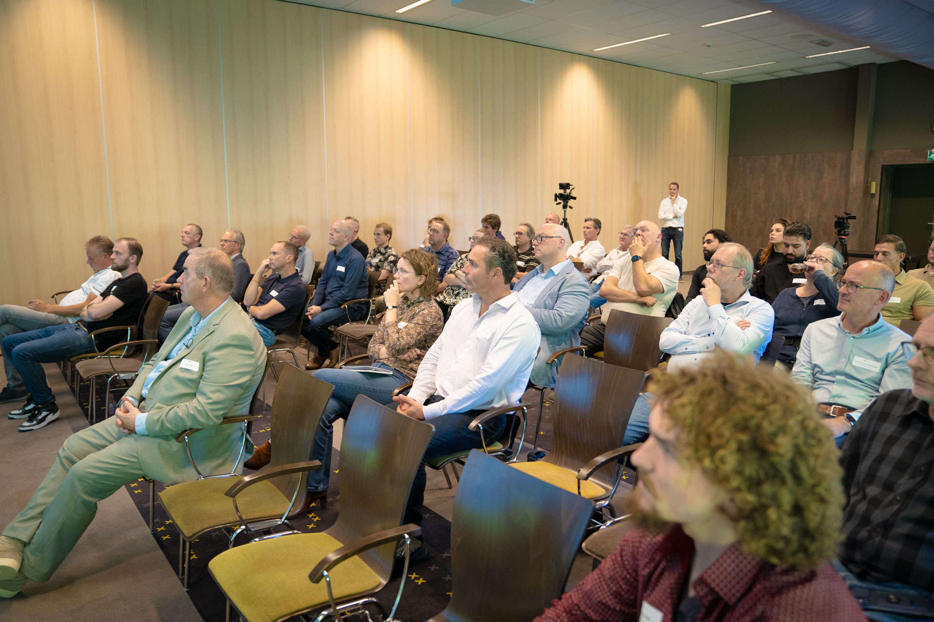 Rows of seated attendees listen attentively in a wood-panelled conference room, with a man standing near the back.