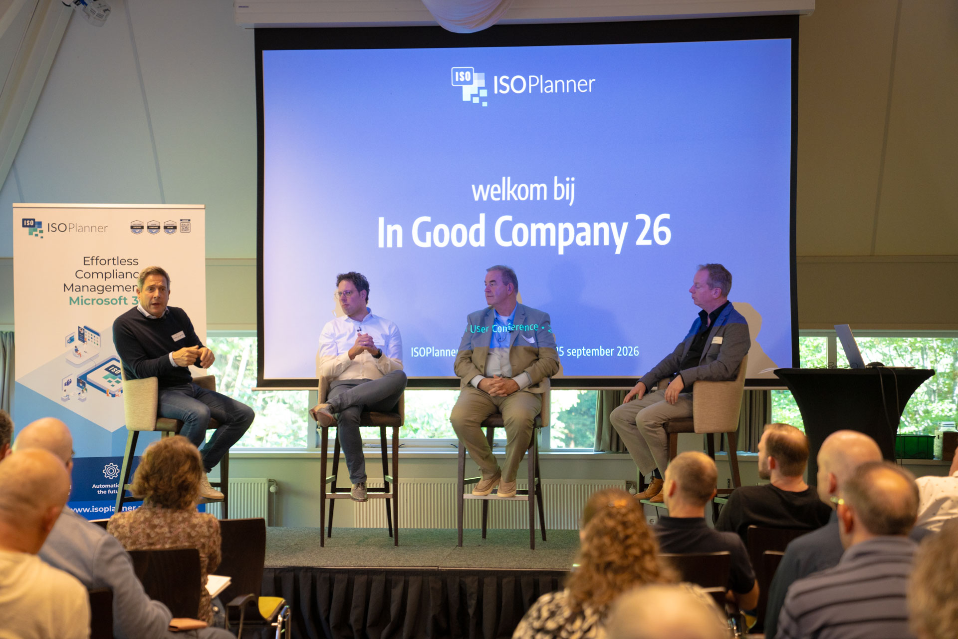 Three men sit on stage stools in front of a welcome screen, with an ISOPlanner™ banner beside them and guests seated below.