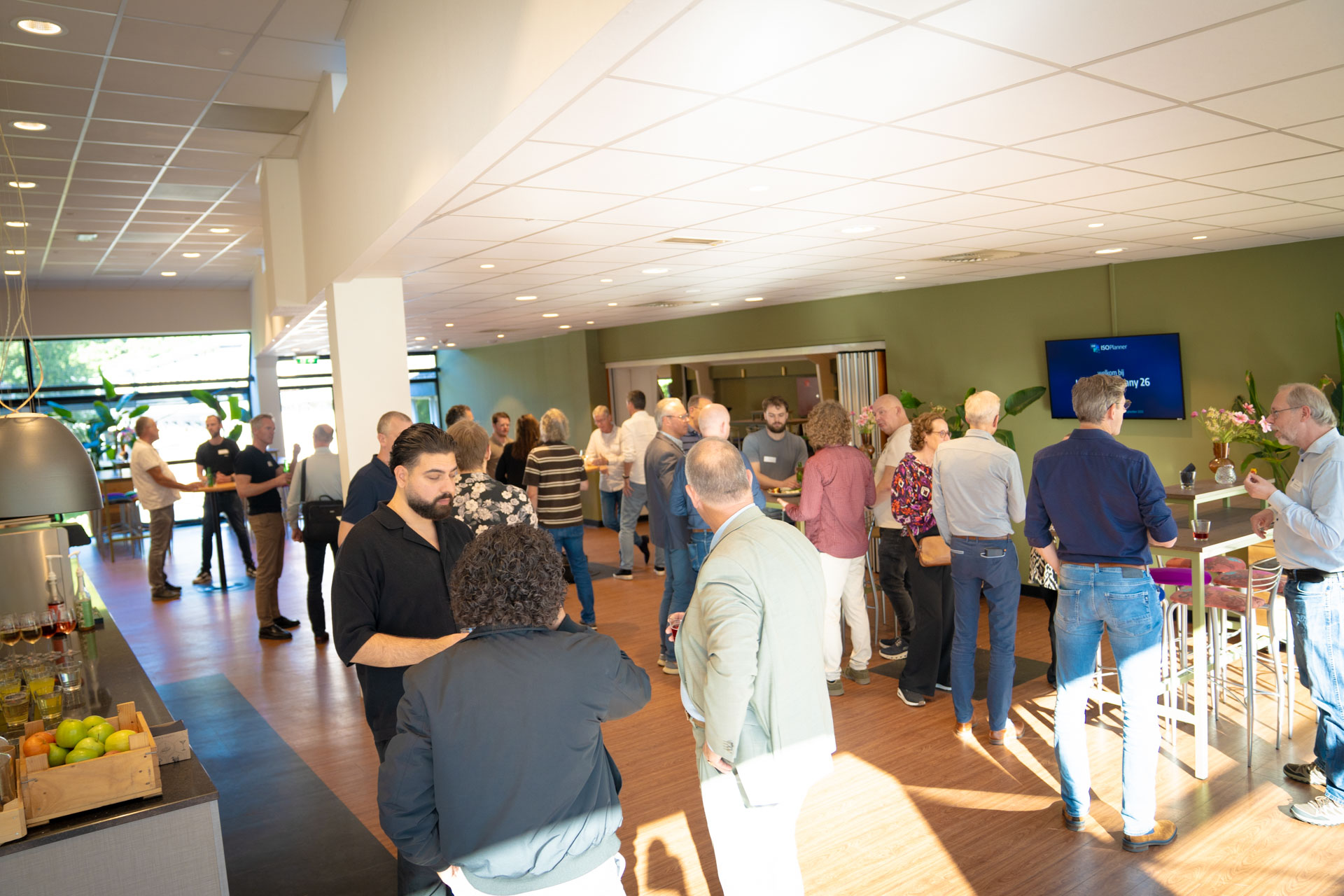 A crowded reception area with around twenty people standing in conversation near a bar, with a welcome screen on the wall.