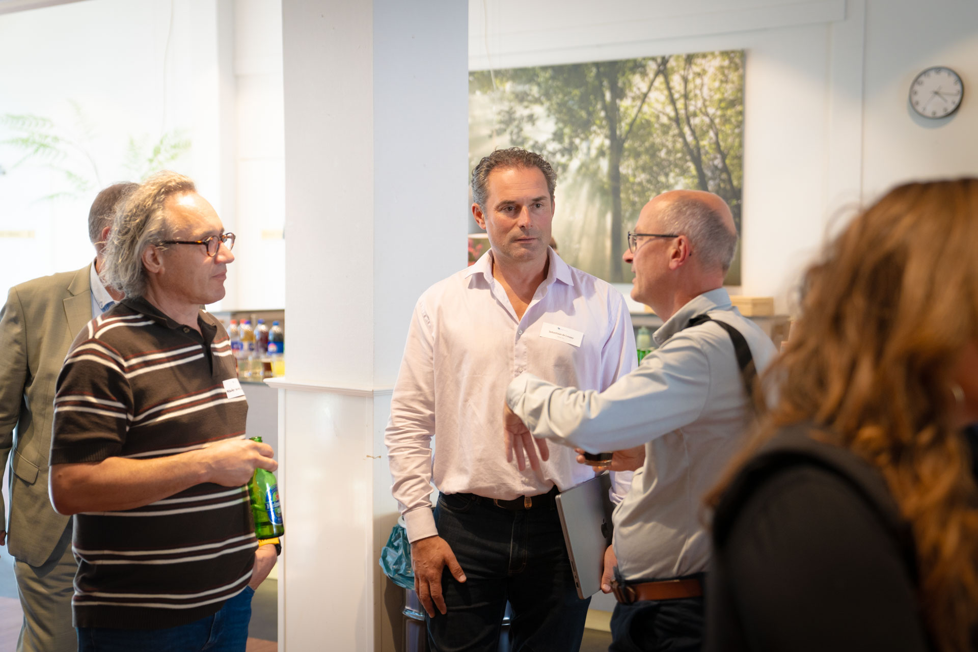 Four men stand talking indoors in front of a large tree photograph, one of them holding a green bottle.