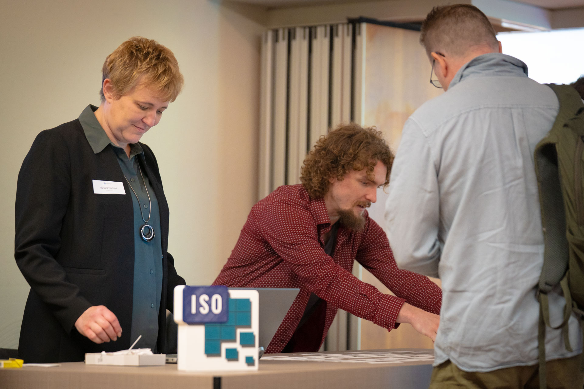 A woman and two men look over papers and a laptop at a registration table marked with an ISO branded sign.