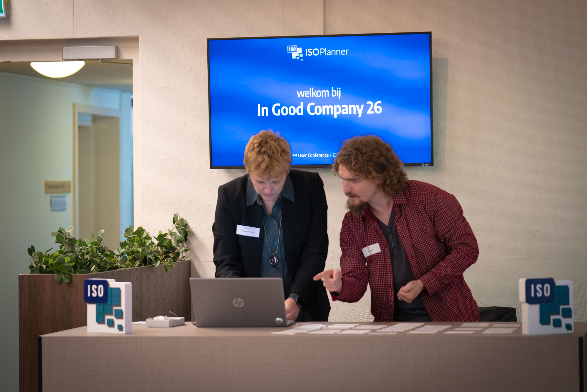 A man points something out on a laptop screen to a woman, standing below the welcome screen for In Good Company 26.
