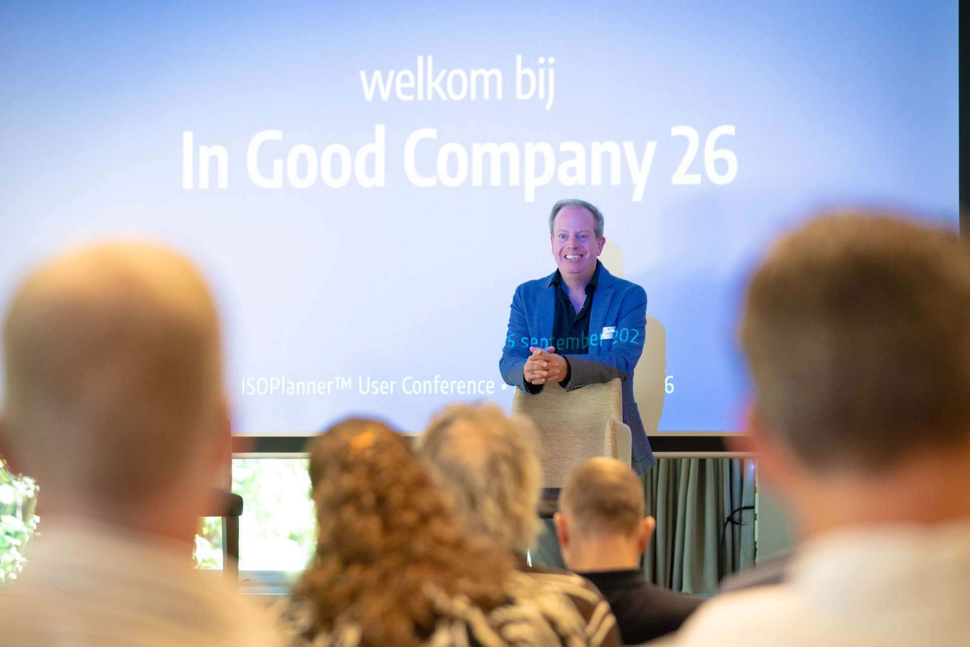 A man stands smiling in front of a screen reading welkom bij In Good Company 26, addressing a seated audience.
