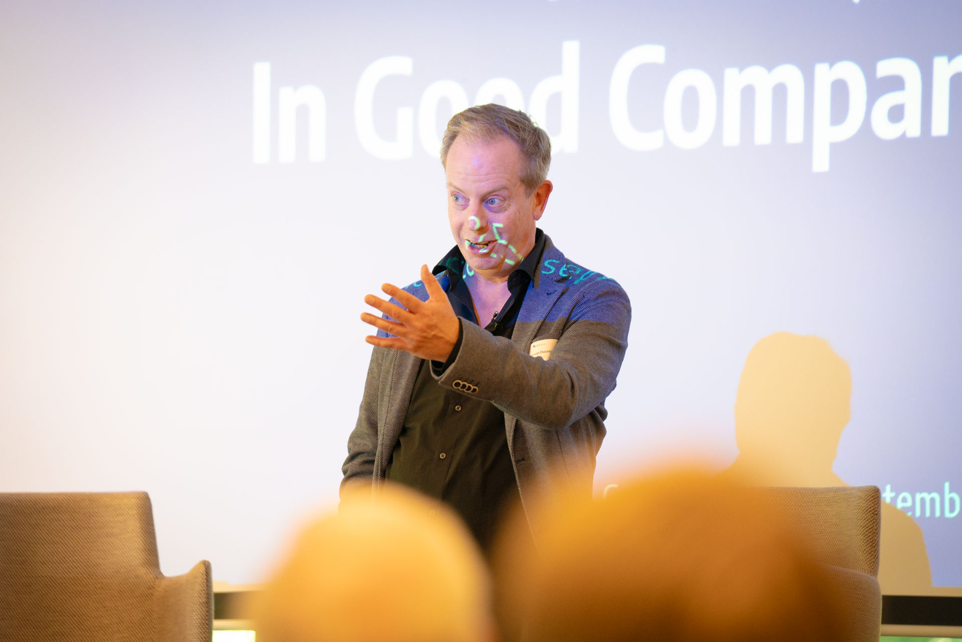 A speaker gestures while addressing the audience, with coloured light effects from the screen falling across him.