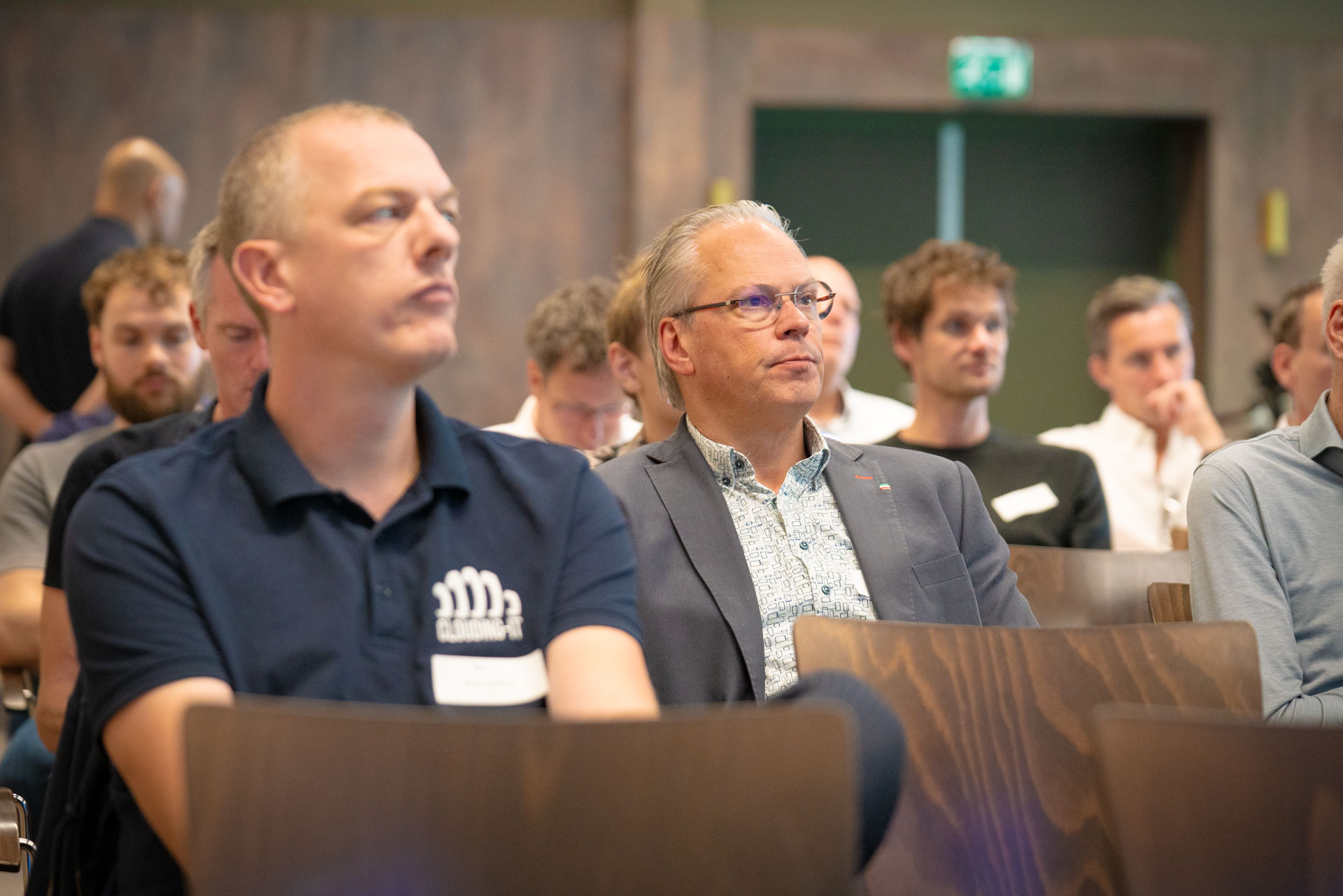 Two seated men watch a presentation, one wearing a polo shirt branded Clouding-IT.