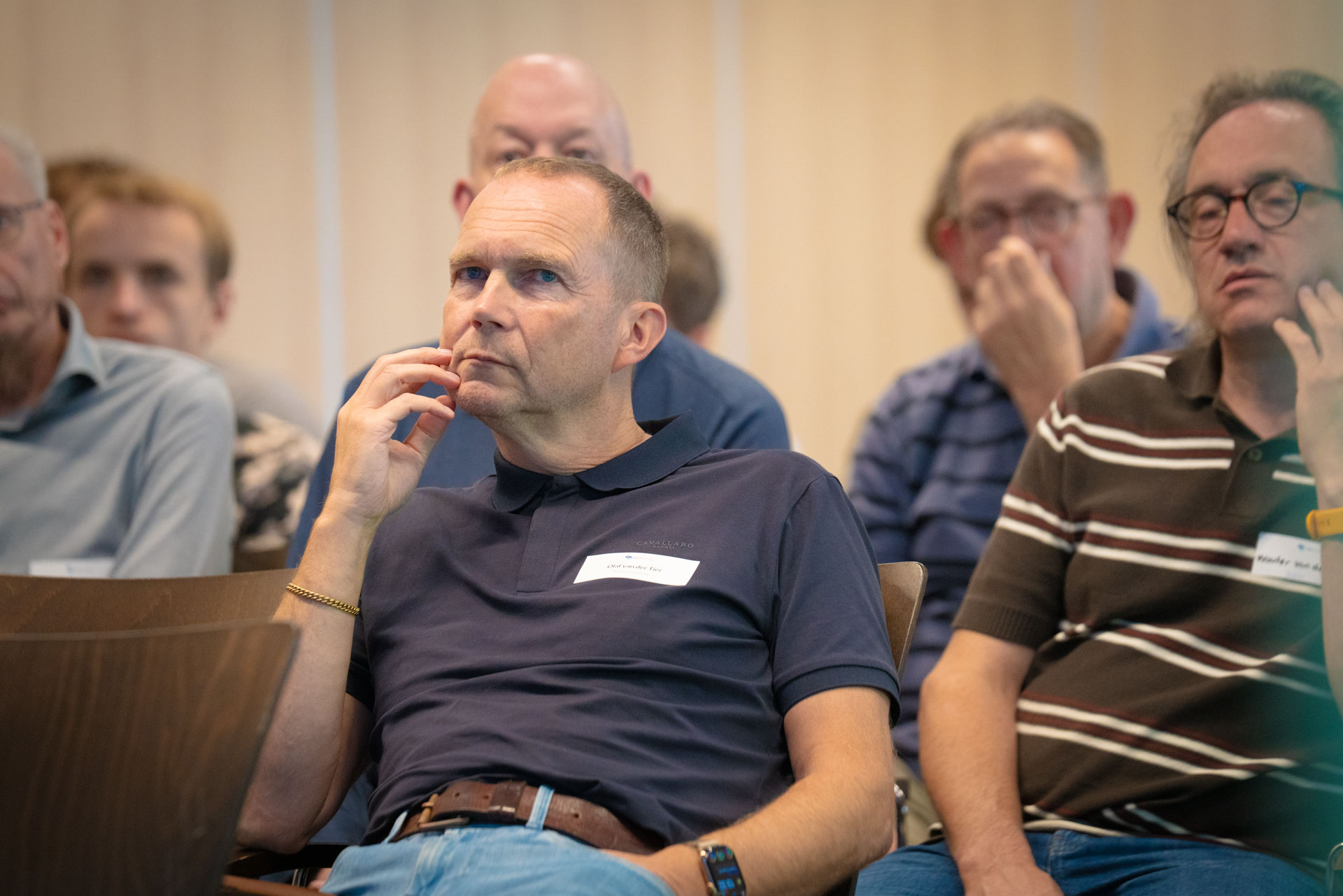 A seated man listens thoughtfully with his hand to his chin, surrounded by other attendees.
