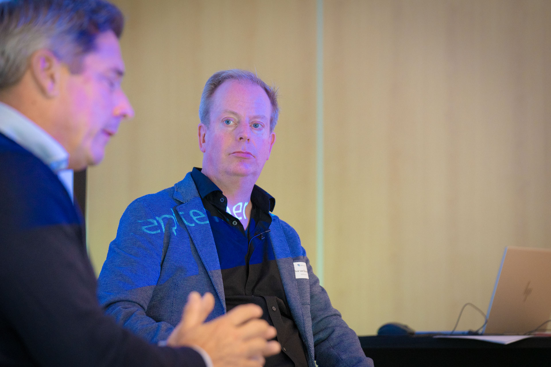 Two men sit on stage in conversation, one lit by green projected text from the screen behind him.