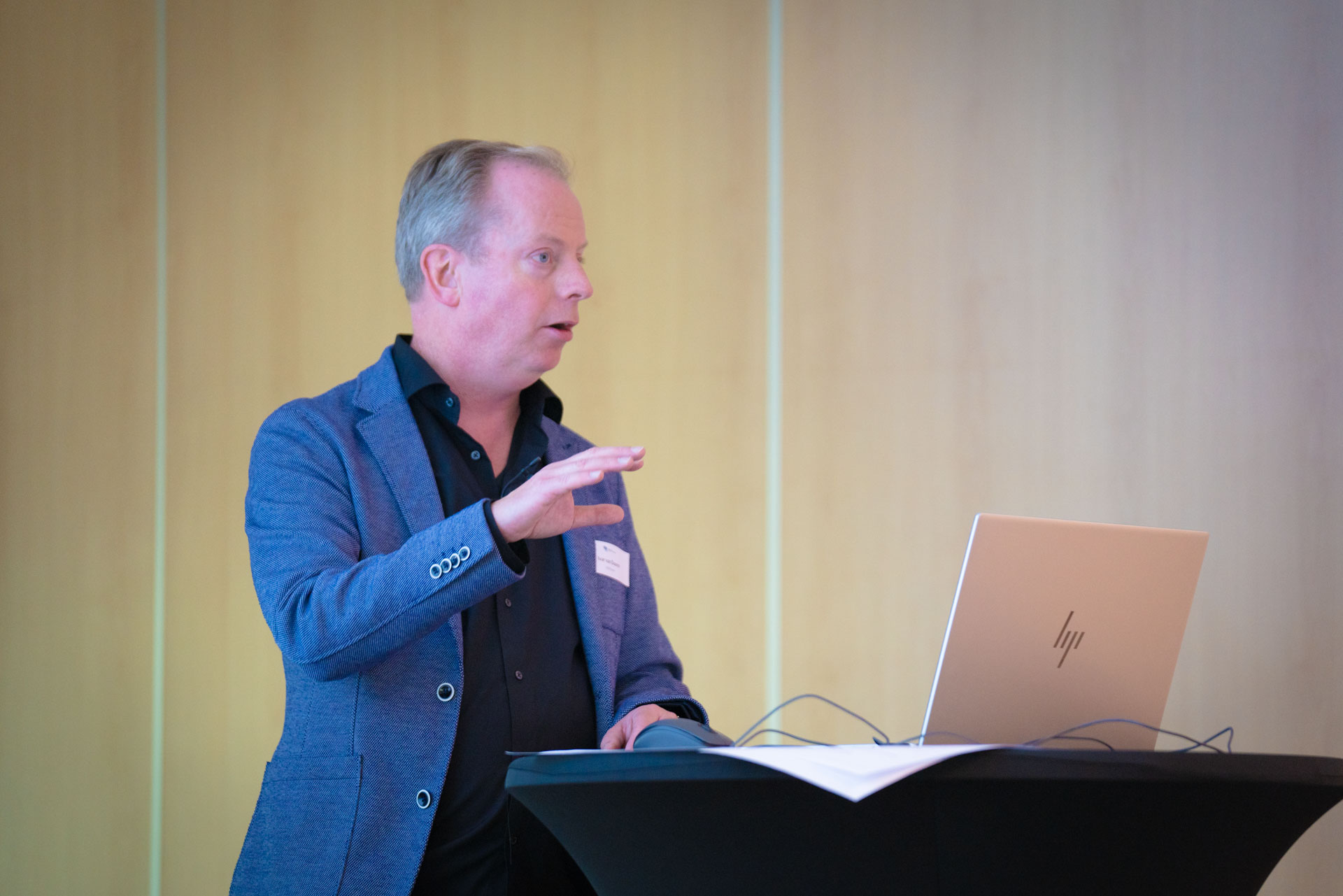 A man speaks at a podium with a laptop open in front of him, gesturing to an unseen audience.