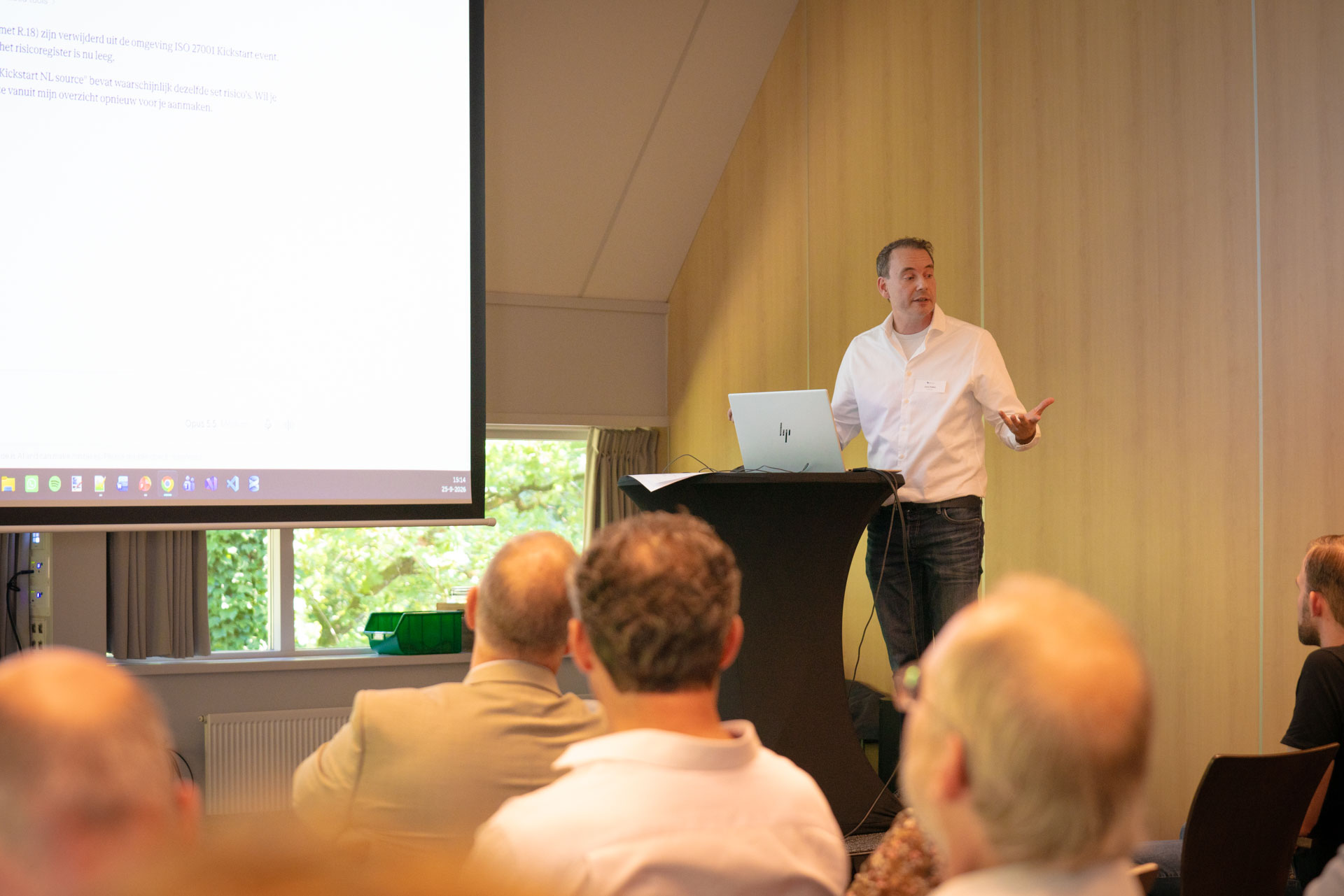 A presenter at a podium shows a slide about the ISO 27001 Kickstart risk register to the audience.
