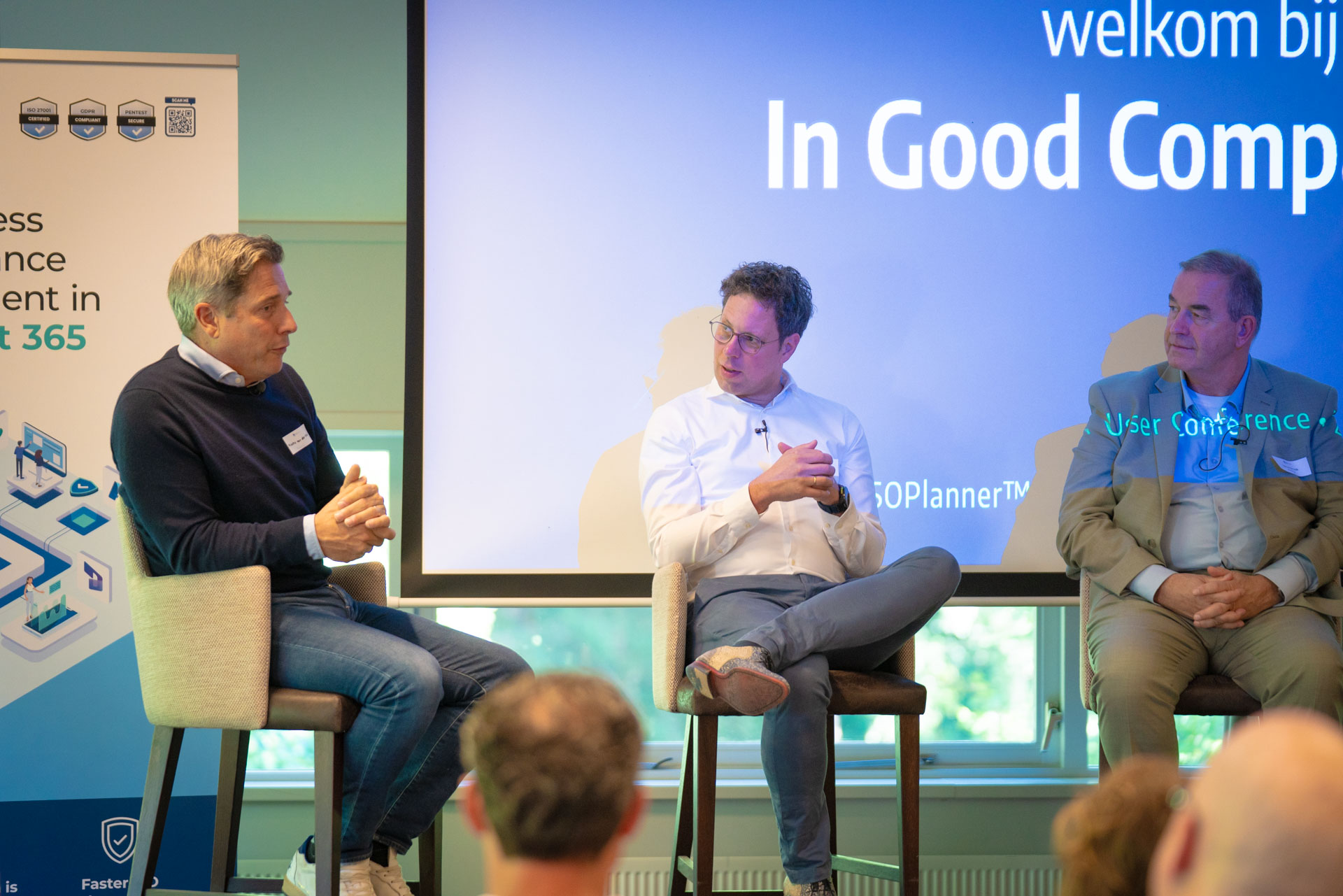Three men sit on stage stools in a panel discussion, with an ISOPlanner™ banner and the audience in the foreground.