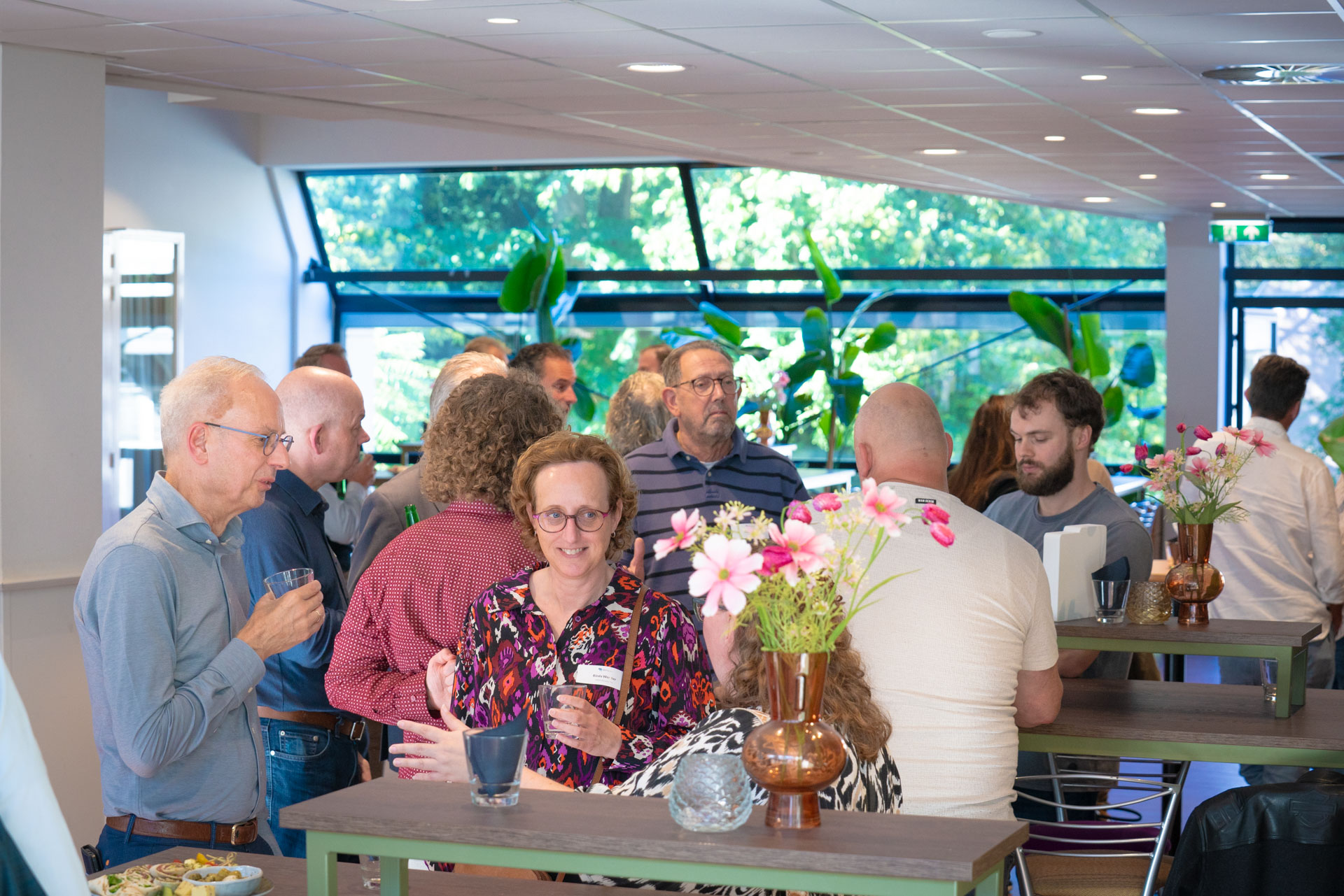 A crowd of guests stand talking and holding drinks near large windows, with flowers on the side tables.
