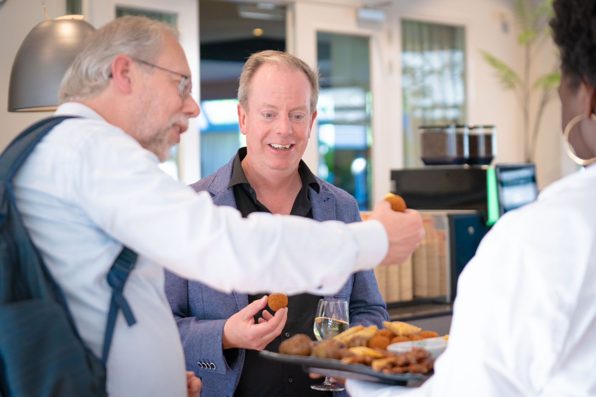 Three people smile while sharing snacks from a tray, with a glass of wine visible on the table.