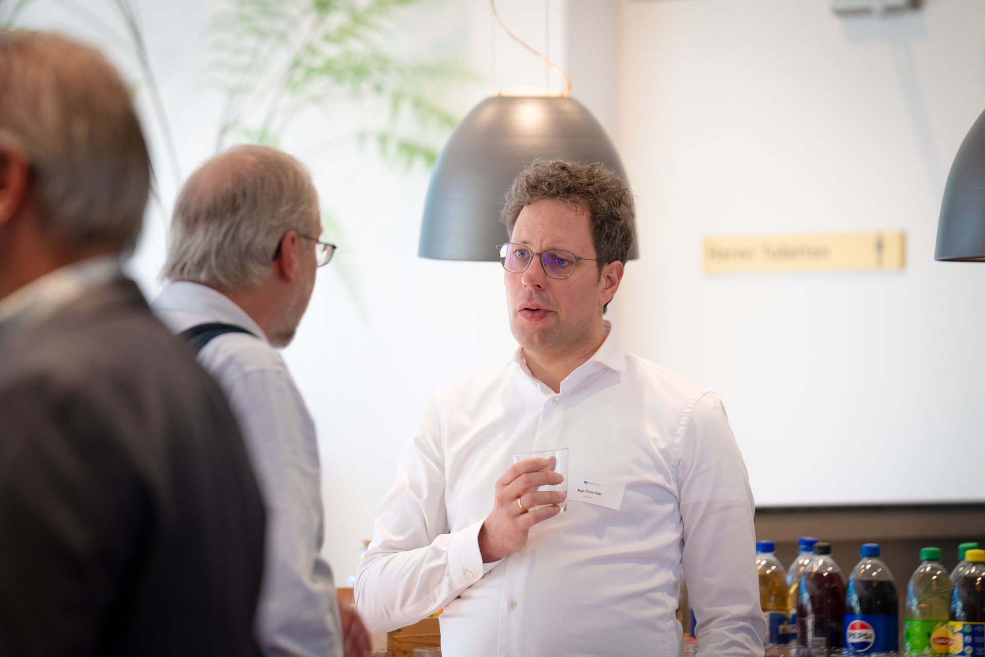 A man talks with two others near the bar area, with pendant lamps and drink bottles visible nearby.