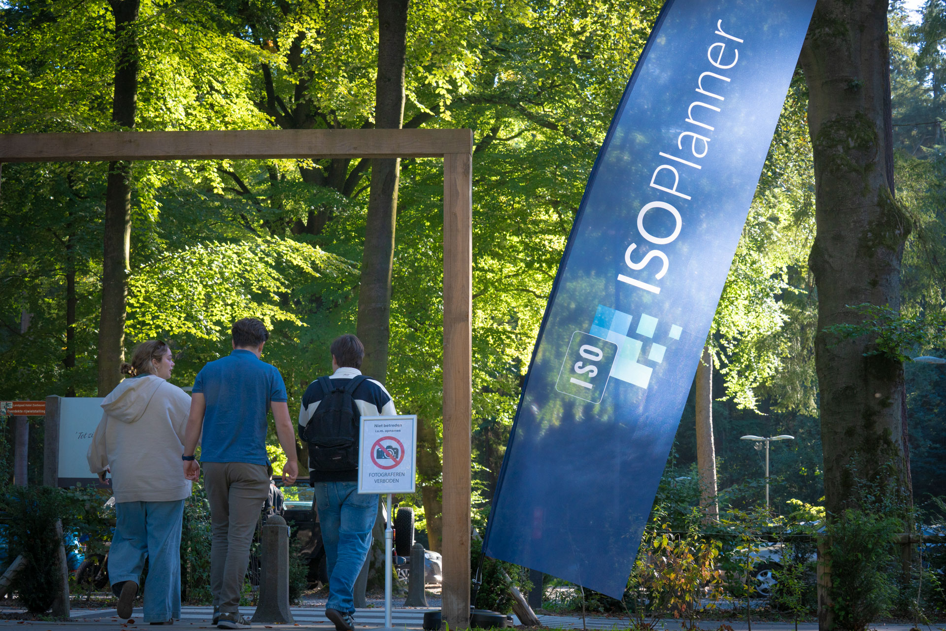 Three people walk away through the forest near the venue, with an ISOPlanner™ flag close in the foreground.