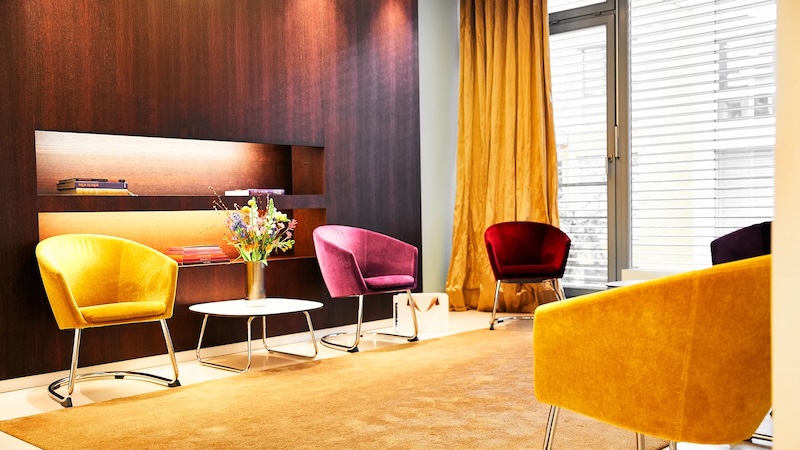 Wartezimmer mit bunten Sesseln, kleinem Tisch und Vase mit Blumen vor Holzwand und Fenster.